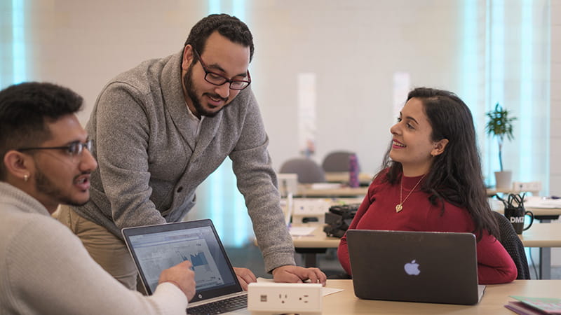 Three employees in conversation at a table with laptops